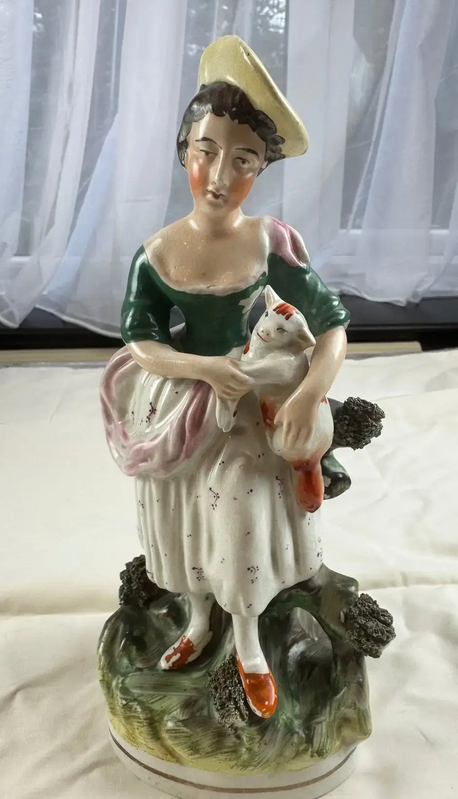 Exquisite 19th-century Staffordshire figurine of a woman with a yellow bonnet cradling a cat amidst lush greenery.